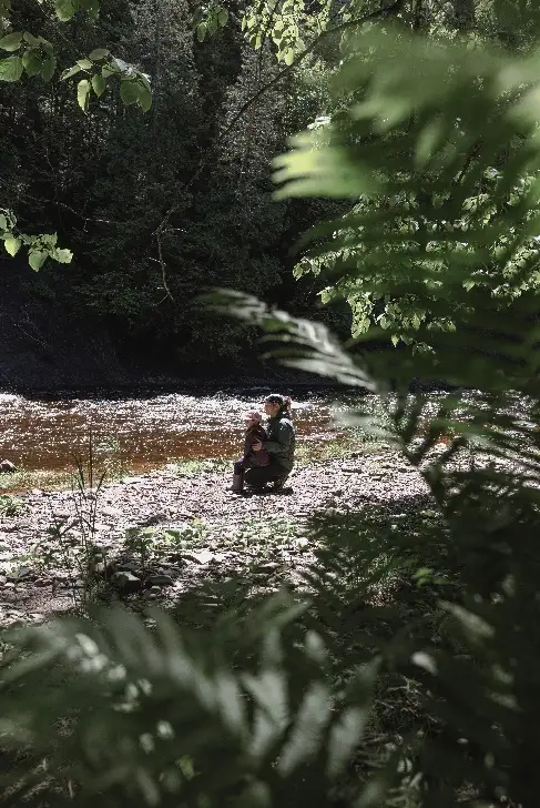 Personne avec enfant près d'une rivière boisée ensoleillée.