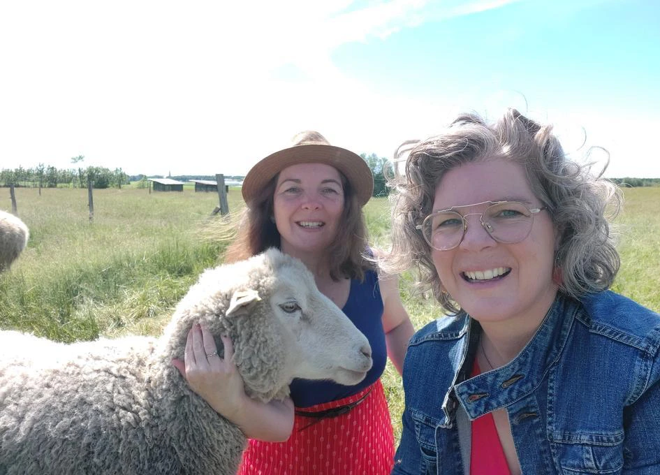 Deux femmes sourient avec un mouton dans un champ.